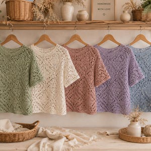 May include: Five short-sleeved shirts made of knit fabric with a repeating diamond pattern, displayed in a row on wooden hangers. The shirts are available in sage green, cream, dusty rose, lavender, and light blue, each featuring a round neckline.