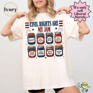 Op de afbeelding: Een ivoorkleurig t-shirt met een afbeelding van acht glazen potten met rood, wit en blauwe deksels en labels. De tekst luidt "CIVIL RIGHTS ARE MY JAM" met labels op de potten zoals "EQUALITY", "DUE PROCESS", "JUSTICE FOR ALL", "FREE SPEECH", "VOTING RIGHTS", "BODILY AUTONOMY", "QUEER RIGHTS" en "ABOLISH ICE".