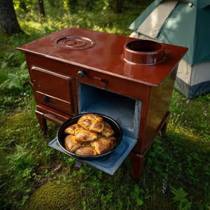 Könnte beinhalten: Ein rostbrauner Outdoor-Ofen mit runder Kochplatte und Backofen. Eine schwarze Schüssel mit Backwaren befindet sich im offenen Ofen. Ein grünes Zelt im Hintergrund deutet auf Camping oder Kochen im Freien hin.