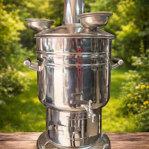 May include: A polished stainless steel samovar, featuring a tall chimney, two small side bowls, and a tap. The tea kettle is placed on a wooden surface with a blurred green backdrop.