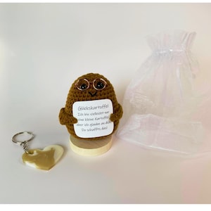 Könnte beinhalten: Eine braune gehäkelte Kartoffelfigur mit schwarzen Augen und Drahtbrille, die eine weiße Karte mit dem Text "Glückskartoffel" hält. Sie steht auf einem Holzsockel neben einem herzförmigen Schlüsselanhänger und einem weißen Organzabeutel.