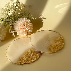 Peut inclure: Deux dessous de verre en résine avec un motif de feuilles d'or. Les dessous de verre sont blanc cassé avec une bordure dorée et un motif de feuilles d'or sur un côté. Les dessous de verre sont ronds avec un bord organique.