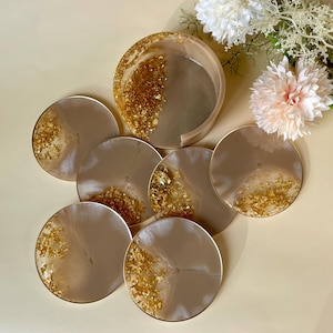Può includere: Un set di sei sottobicchieri rotondi e un porta bicchieri. I sottobicchieri e il porta bicchieri sono beige con accenti di foglie d'oro. I sottobicchieri sono disposti a cerchio. Il porta bicchieri è cilindrico. Lo sfondo è beige chiaro.