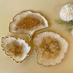 Peut inclure: Trois plateaux décoratifs en forme de feuilles avec des bordures dorées. Les plateaux sont blancs avec des paillettes dorées incrustées dans la résine. L'un des plateaux a la forme d'une feuille de ginkgo, les deux autres ont la forme d'une feuille. Un bracelet repose sur un plateau.