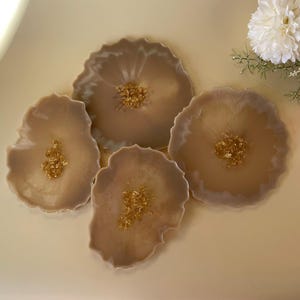 Puede incluir: Cuatro posavasos de resina beige con motas doradas dispuestos sobre una superficie beige claro. Los posavasos tienen una forma natural y orgánica. Flores blancas y rosas en el fondo.