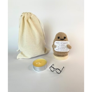 Könnte beinhalten: Eine beige gehäkelte Kartoffelfigur, die ein Schild mit der Aufschrift "Emotionale Support-Kartoffel Meine Aufgabe? Einfach da sein. Manchmal reicht das schon." hält. Das Set enthält ein gelbes Teelicht, eine schwarze Miniaturbrille und einen Beutel.