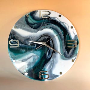 May include: A round wall clock featuring a swirling abstract pattern in shades of deep teal, dark blue, white, and silver. The clock face has metallic silver numerals at the 12, 3, 6, and 9 o'clock positions, with simple silver hands at the center.