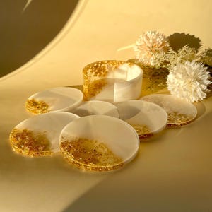 Op de afbeelding: Een set witte en gouden hars onderzetters en een bijpassende houder. De onderzetters zijn rond met goudbladaccenten. De houder heeft een cilindrische vorm met goudblad details. De items zijn geplaatst op een gele ondergrond.