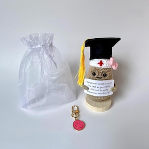 Könnte beinhalten: Eine gehäkelte beige Figur mit schwarzem Doktorhut und gelber Quaste, einer weißen Krankenschwesterhaube mit rotem Kreuz und einer Drahtbrille. Sie hält ein Schild mit der Aufschrift "Herzlichen Glückwunsch! Du hast es geschafft. Die Welt braucht Menschen wie dich." Ein rosa Smiley-Schlüsselanhänger und ein weißer Organzabeutel liegen bei.