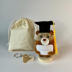 Könnte beinhalten: Ein gehäkelter brauner Bär mit schwarzer Doktorhut-Kopfbedeckung, gelber Quaste und Drahtbrille. Der Bär hält ein weißes Schild mit der Aufschrift "Personlisiere mich!" und steht auf einem Holzsockel neben einem herzförmigen Schlüsselanhänger und einem Beutel.