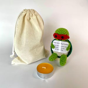 Könnte beinhalten: Eine gehäkelte Schildkröte mit roter Maske hält ein weißes Schild mit der Aufschrift Little Turtle, Big Dreams, Große Ziele brauchen Zeit. Glaube an dich und geh deinen Weg. Dazu ein beiger Kordelzugbeutel und ein rundes gelbes Teelicht.