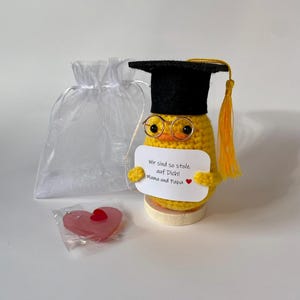 Op de afbeelding: Een geel gehaakt kuikentje met een zwarte afstudeerhoed met gele kwast en een goudkleurige bril. Het houdt een wit bordje vast met de tekst "Wir sind so stolz auf Dich! Mama und Papa" en een rood hartje. Inclusief een wit zakje en een roze hartje.