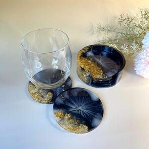May include: A clear glass tumbler sits on a round black and white resin coaster with gold leaf accents. Beside it, a matching coaster lies flat, and a black resin coaster holder contains two additional coasters with similar gold leaf detailing.