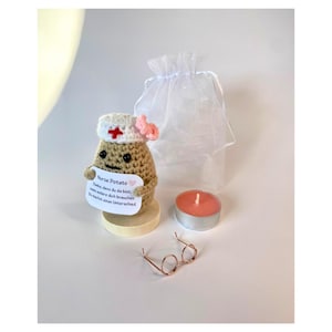 Op de afbeelding: Een gehaakt beige aardappelpoppetje met een wit verpleegsterskapje met een rood kruis en een roze strik. Het houdt een wit bordje vast met de tekst Nurse Potato, Danke, dass du da bist, wenn andere dich brauchen, Du machst einen Unterschied. Inclusief een waxinelichtje, een brilletje van metaaldraad en een wit organza zakje.