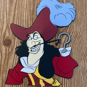 May include: A Captain Hook cutout, featuring a red coat, black hair, and a white shirt. The character has a sinister grin and a silver hook. A light blue feather plume adorns his hat. The cutout is mounted on a white stick.