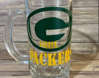 May include: A clear glass beer mug with a handle. The mug features a green and yellow design of the Green Bay Packers logo and team name. The mug is made of clear glass and has a textured base.