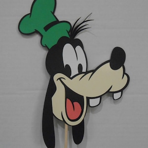 Puede incluir: Un recorte de Goofy, un personaje de dibujos animados, con un sombrero verde, pelaje negro y un gran hocico color crema. El personaje sonríe, revelando una lengua roja y dos dientes blancos. Se adjunta un palo de madera.