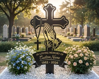May include: A black metal memorial cross featuring a cutout design of praying hands and a hanging rosary. A banner at the base displays the text IN LOVING MEMORY OF Custom Name 1962 - 2026. The cross is positioned in a cemetery setting.