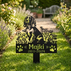 以下が含まれることがあります： 犬のシルエットと花の切り抜きが施された黒い金属製のガーデンステーク。サインには「Majki」と「2011-2024」の年号と足跡が書かれています。犬はコッカー・スパニエルです。庭に置かれたステークです。