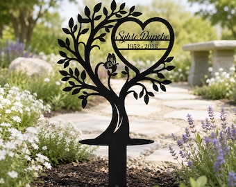 May include: Black metal garden stake in the shape of a tree with a heart-shaped cutout. The heart contains the name "Sylvie Dupêbe" and the years "1952-2018". A butterfly is perched on a branch. The stake is in a garden setting.