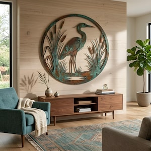 May include: A circular metal wall art piece featuring a heron standing among cattails. The design has a weathered, oxidized copper finish with shades of teal, green, and brown. The artwork is framed within a thin circular metal border.
