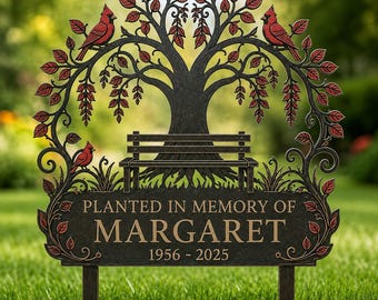 May include: A dark metal memorial garden stake featuring a tree with red leaves and three red cardinal birds. A bench sits at the base of the tree above a rectangular plaque with the text PLANTED IN MEMORY OF MARGARET 1956 - 2025.