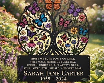 May include: A black metal garden stake featuring a circular tree design with colorful butterflies and flowers. Below the tree, a rectangular plaque displays the text: THOSE WE LOVE DON'T GO AWAY, THEY WALK BESIDE US EVERY DAY. SARAH JANE CARTER 1955 - 2024.
