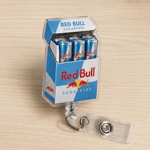 K&ouml;nnte beinhalten: Ein ausziehbarer Ausweishalter mit einem rechteckigen Miniatur-Schachteldesign, das das Red Bull Sugarfree Logo und Branding tr&auml;gt. Die Schachtel enth&auml;lt f&uuml;nf kleine, zylindrische silber-blaue Dosen mit Red Bull Branding und einen transparenten Clip.