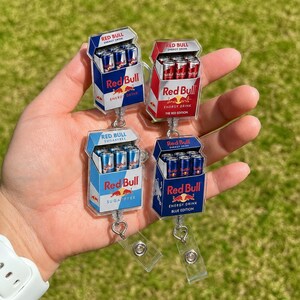 K&ouml;nnte beinhalten: Vier Acryl-Ausweishalter mit Miniatur-Red-Bull-Energy-Drink-Kartons. Die Designs umfassen die originale blaue Version, Red Edition, Sugarfree und Blue Edition, jeweils mit dem Red-Bull-Logo und Schriftzug auf einer transparenten Basis.