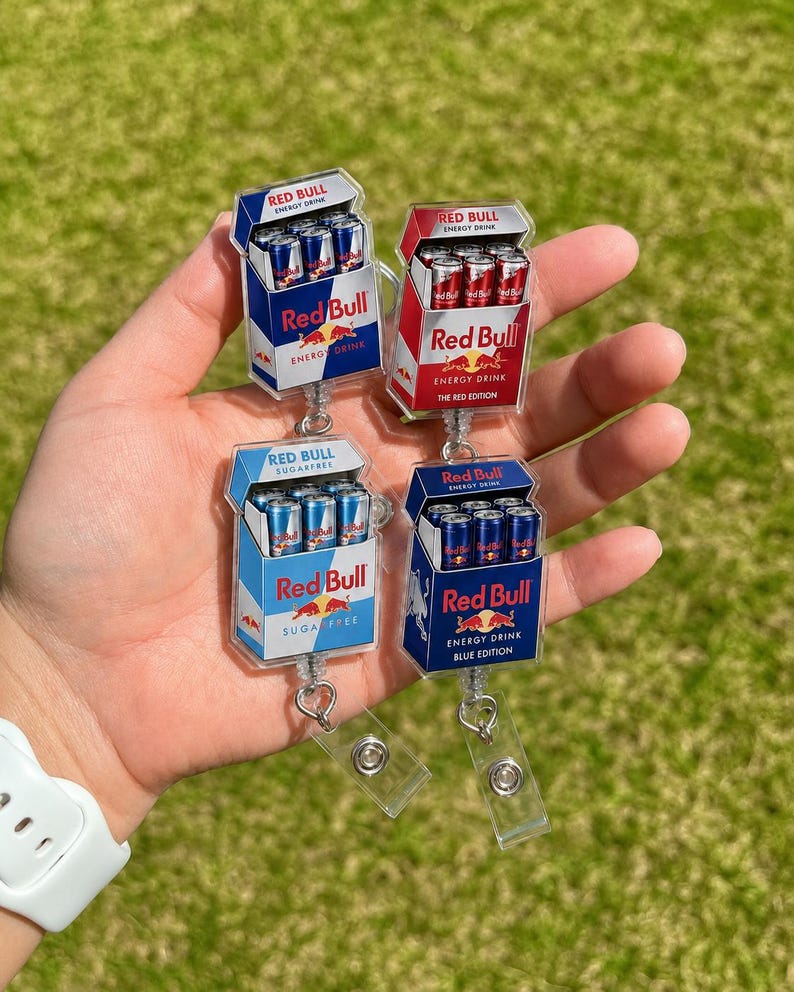 K&ouml;nnte beinhalten: Vier Acryl-Ausweishalter mit Miniatur-Red-Bull-Energy-Drink-Kartons. Die Designs umfassen die originale blaue Version, Red Edition, Sugarfree und Blue Edition, jeweils mit dem Red-Bull-Logo und Schriftzug auf einer transparenten Basis.