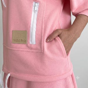 May include: A light pink fleece sweatshirt featuring a white zippered pocket and a rectangular tan patch with the text Wild Pelt and a cat illustration. The garment includes a white drawstring at the hem and a side pocket opening.