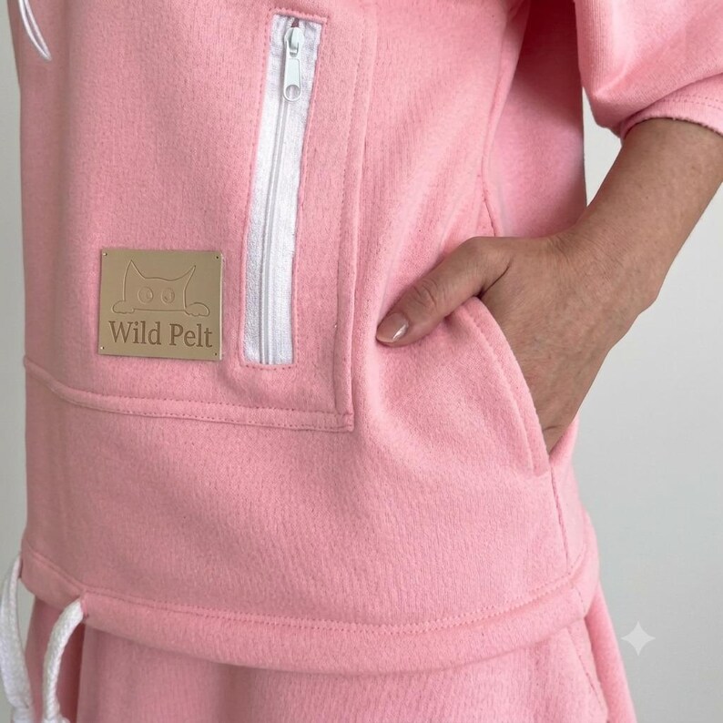 May include: A light pink fleece sweatshirt featuring a white zippered pocket and a rectangular tan patch with the text Wild Pelt and a cat illustration. The garment includes a white drawstring at the hem and a side pocket opening.