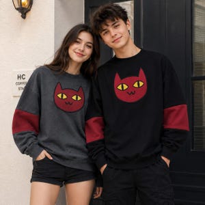 May include: A charcoal grey sweatshirt and a black sweatshirt, both featuring a red cat face graphic with yellow eyes and a small mouth. Each long-sleeved garment has a contrasting red band on the lower sleeves and a crew neckline.