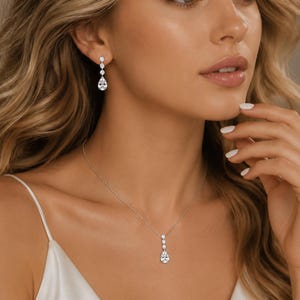 May include: A silver-toned jewelry set consisting of a necklace and a pair of drop earrings. Each piece features a vertical arrangement of two round clear stones topped by a single pear-shaped clear stone, all set in a polished metallic frame.