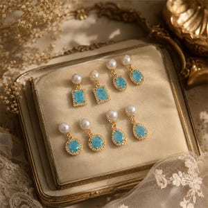 May include: A collection of gold-toned earrings with various shapes, including squares, ovals, circles, and teardrops. Each earring features a light blue stone, surrounded by small clear crystals, and a dangling white pearl. The earrings are displayed on a cream-colored surface.