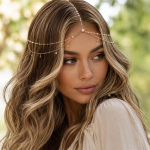 May include: A gold-toned headpiece featuring a delicate chain design that rests across the forehead. The accessory includes a central circular stone and multiple smaller dangling crystal accents suspended from two thin, layered metallic chains.