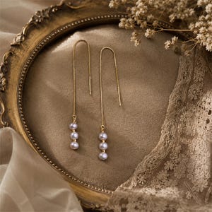 May include: A pair of gold-colored threader earrings with three light purple pearls dangling from each earring. The earrings are displayed on a gold-framed, oval-shaped tray with a neutral-colored background.