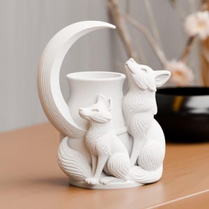 May include: A matte white ceramic vessel featuring a sculpted crescent moon and two foxes. One fox sits at the base while the other stands beside it, looking upward. The cylindrical container is integrated into the center of the moon and fox design.