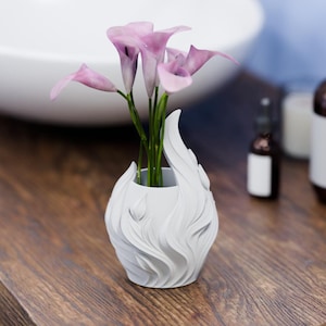 Pode incluir: Um vaso branco com uma textura exterior esculpida em forma de chama contendo um ramo de lírios calla roxo-claros com caules verdes. O vaso tem uma base arredondada e uma borda assimétrica e pontiaguda que imita chamas a subir.