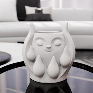May include: A matte white ceramic planter featuring a sculpted face with closed eyes and a subtle smile. The exterior is decorated with raised, teardrop-shaped elements surrounding the central face, creating a textured and dimensional appearance.