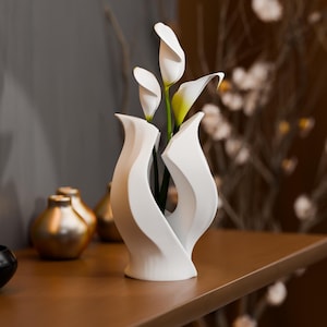 Op de afbeelding: Een witte keramische vaas met een gladde, gebogen, abstracte vormgeving met drie witte calla lelies met groene stelen. De vaas heeft een gespleten ontwerp dat een open midden creëert, waardoor de donkergroene stelen zichtbaar zijn.