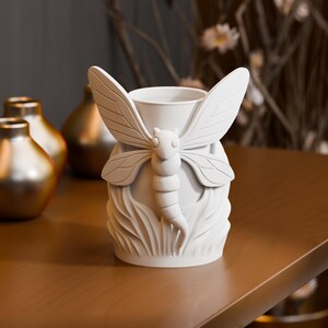 Könnte beinhalten: Eine matte, weiße zylindrische Vase mit einem Relief einer Libelle mit ausgebreiteten Flügeln in der Mitte. Der Sockel der Vase ist mit vertikalen, blattartigen Mustern strukturiert, die den unteren Körper des Insekts umgeben.