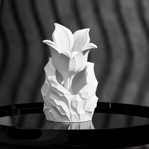 Könnte beinhalten: Ein mattweißes skulpturales Objekt mit einer zentralen, blühenden Blumenform, die aus einer Basis mit facettierten, felsartigen geometrischen Texturen hervorgeht. Das Stück hat eine glatte, gleichmäßige Oberfläche und ein symmetrisches Design.