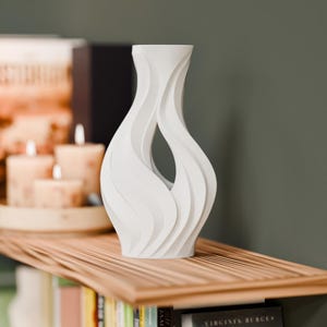 May include: A matte white vase featuring a sculptural, fluid design with vertical, wave-like ridges and a central open aperture. The vase has a narrow neck that flares slightly at the top and a wider, rounded base, creating a continuous, organic form.