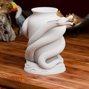 Puede incluir: Un jarrón de cerámica blanco mate con un dragón esculpido envuelto alrededor del exterior. El dragón tiene un cuerpo texturizado y escamoso con la boca abierta y dientes visibles. La base del jarrón presenta un diseño ondulado.