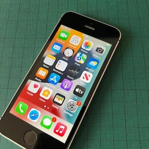 Könnte beinhalten: Ein schwarzes Smartphone mit einem weißen Rand um den Bildschirm, das einen Startbildschirm mit verschiedenen bunten App-Symbolen anzeigt, darunter FaceTime, Kalender, Fotos, Kamera, Mail, Uhr, Karten, Wetter, Notizen, Erinnerungen, Aktien, Bücher, App Store, Podcasts, TV, Gesundheit, Home, Wallet und Einstellungen.