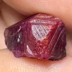 Puede incluir: Una piedra preciosa en bruto y sin pulir con un tono magenta intenso y rojo oscuro. La piedra presenta una superficie irregular y multifacética con texturas cristalinas visibles, estrías naturales y una calidad semitranslúcida en su estructura.