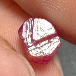 May include: A small, raw, deep pinkish-red gemstone with a faceted, irregular shape. The surface displays prominent, linear striations and reflective, metallic-looking white inclusions that create a high-contrast, textured appearance across the stone.