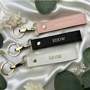 Pode incluir: Três chaveiros com ferragens douradas e texto gravado. Um é rosa com "JOHANNA", um é preto com "EDOW" e um é branco com "BRIDE". Os chaveiros estão sobre uma superfície de cetim branco com vegetação.