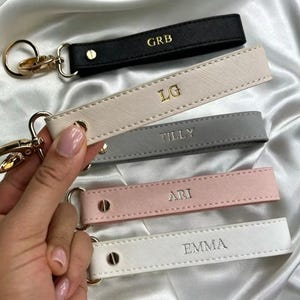 Può includere: Una collezione di portachiavi personalizzati in vari colori: nero, beige, grigio, rosa e bianco. Ogni portachiavi presenta un fermaglio color oro ed è impresso con un nome o iniziali in un colore a contrasto. I portachiavi sono esposti su una superficie bianca e setosa.